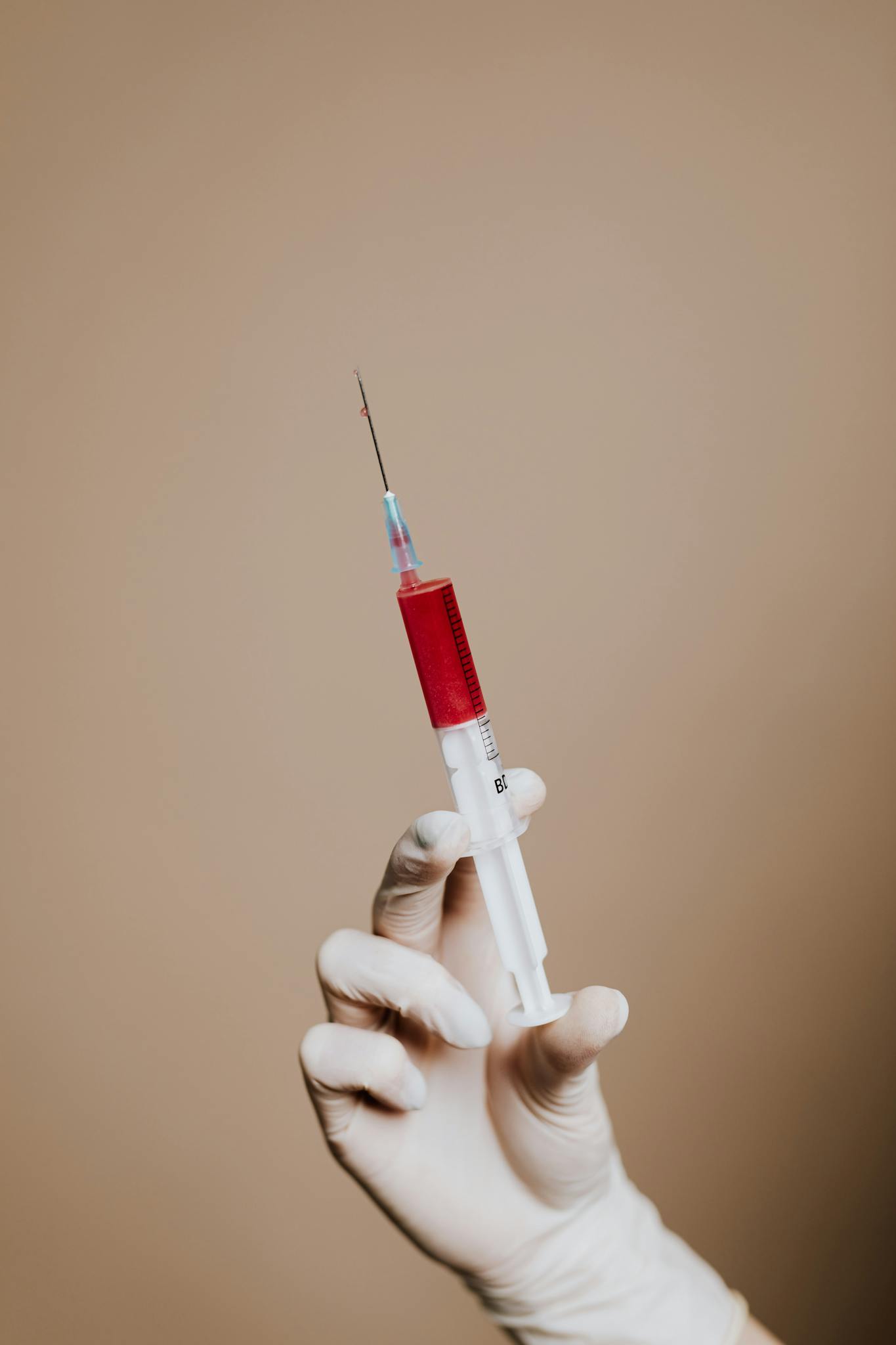 A close-up of a gloved hand holding a syringe filled with red liquid against a neutral background.