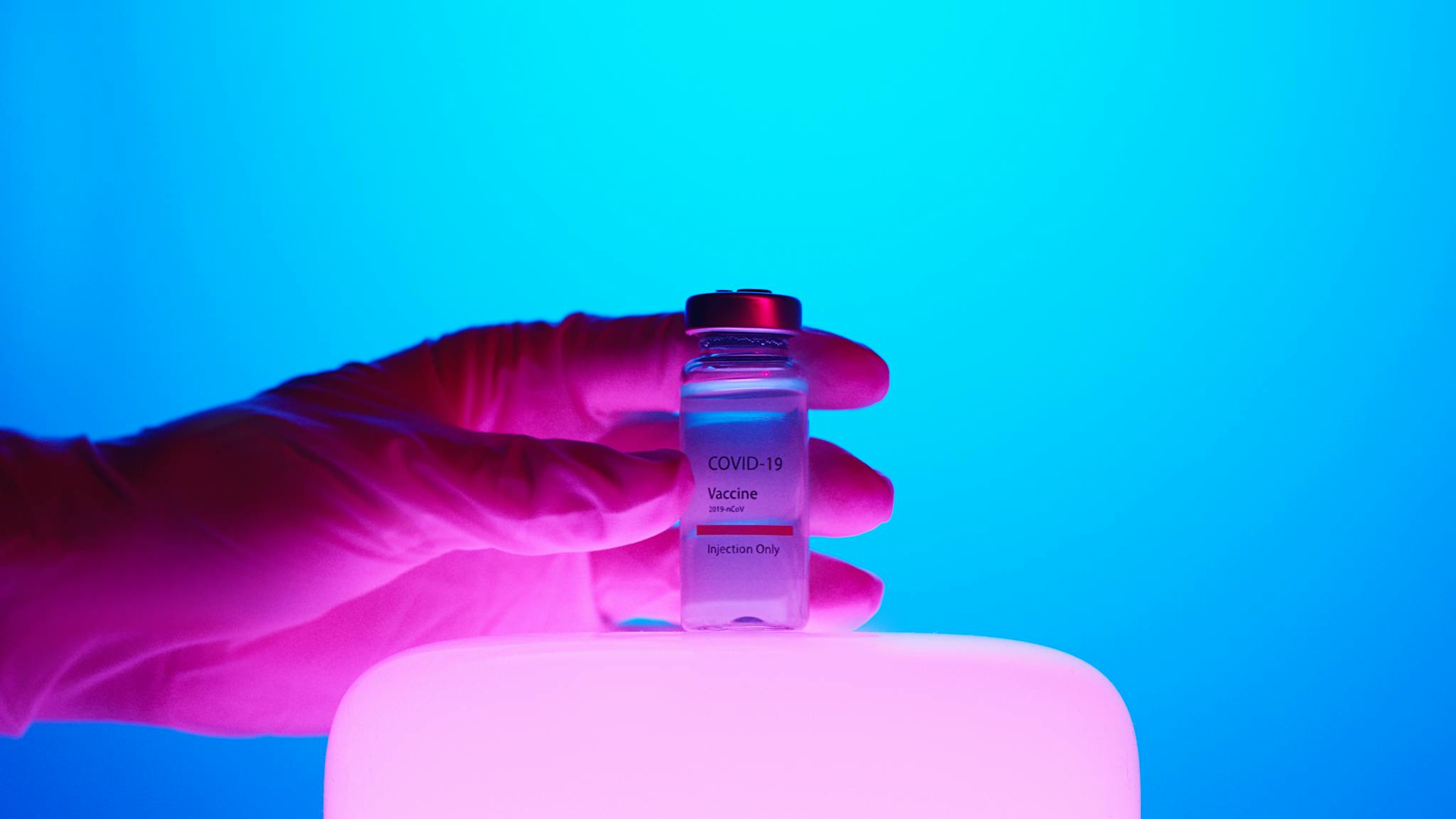 Hand holding a COVID-19 vaccine vial against a vibrant blue background.